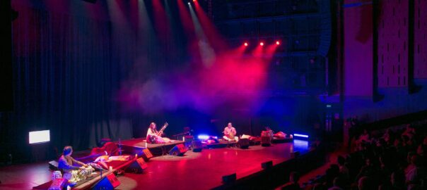 The full band on stage, seated on low risers, with the auditorium lit in blue and read, with haze in the air. To the right of the picture you can see the audience, enraptured