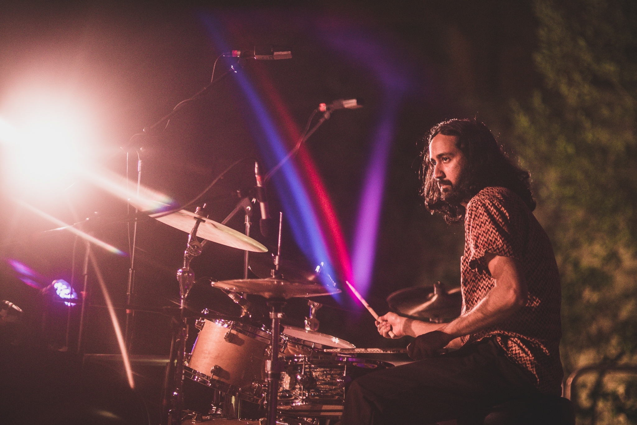 Sarahty Korwar sits at his drumkit, on ethe right hand side of the shot, looking left. The stage is lit with purple and maroon, and has curved brick wall behind - perhaps a railway arch or similar.