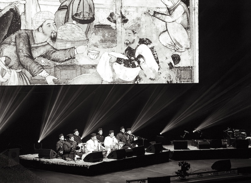Black and white image of the full qawwal band on stage, seated on a low riser, with a large screen behind them, onto which pictures are being projected.
