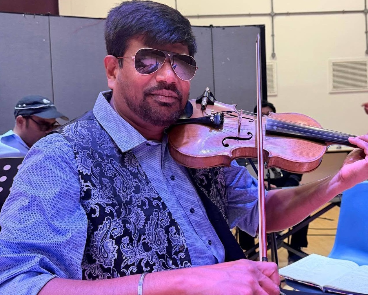 The violinist wearing a blue shirt and floral patterned waistcoat, plays his violin. the photp looks to have been taken at a rehearsal