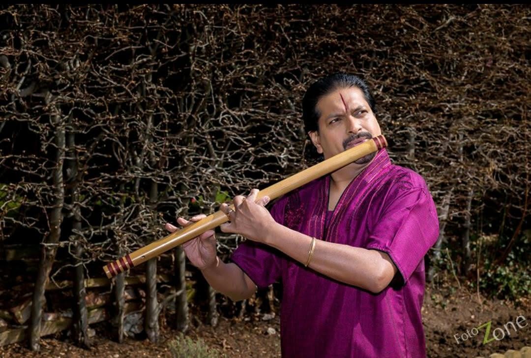 The flautist, platying his flute, and wearing a purple shirt. The shot is a promotional one, taken outside, with foliage behind.